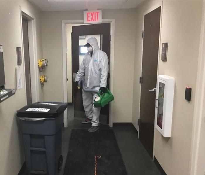 SERVPRO crew member in white PPE is in commercial offce building with green servproxide fogger helping to spray for COVID-19